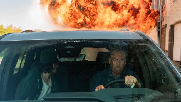 Bob Odenkirk drives away by car in "Nobody 2", while fiery explosion clouds get into the air behind him