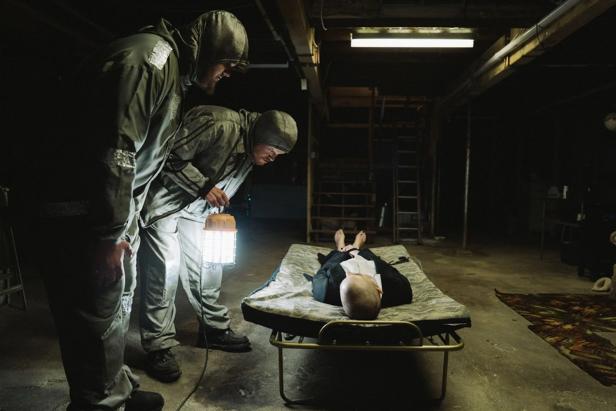 Two men in protective suits lean over a woman on a lounger in a dark basement