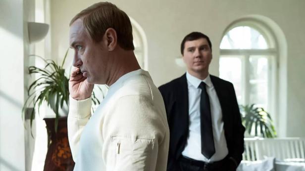 Jude Law as Putin is standing on the phone at the window - in the background is Paul Dano as Baranov
