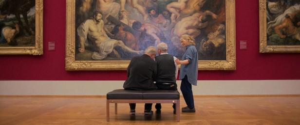 Two men sit on a bench in front of a painting while a woman stands next to them.