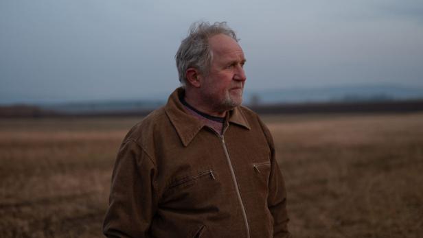 An older man with gray hair stands in a vast, autumnal landscape and looks to the right.