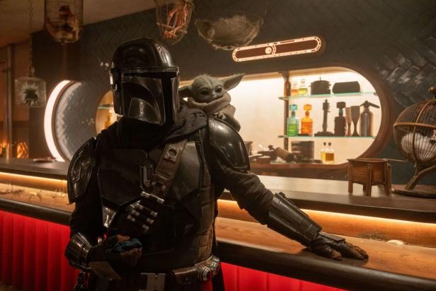 The Mandalorian with Grogu on his shoulder leans against a bar.