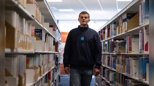 A man wearing a blue necklace stands between bookshelves in a library.