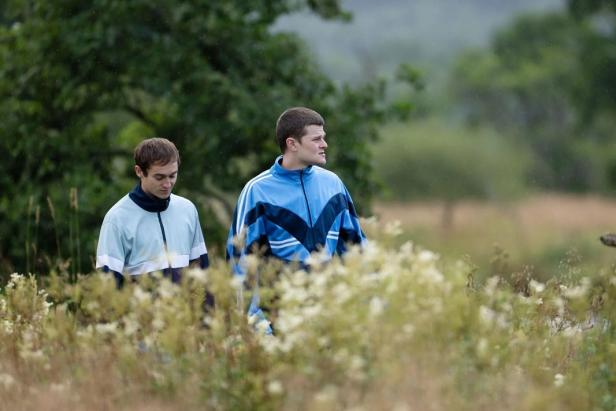 Two young men in training jackets stand in a meadow with wildflowers, trees and blurred nature in the background.