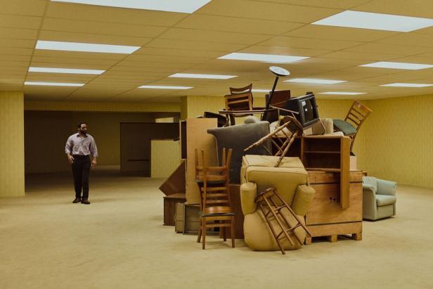 A man stands in a yellow-lit room with a large pile of stacked furniture and objects.