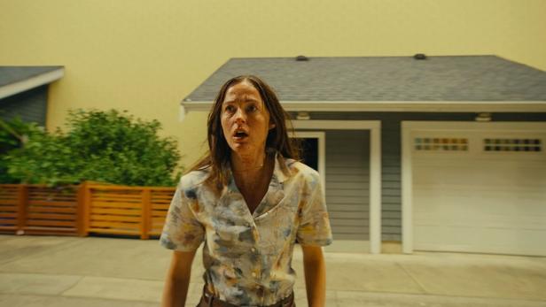 A woman with a dirty face and shirt stands scared in front of a house with a garage and a wooden fence.