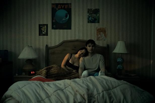 A young couple sits on a bed in a dimly lit bedroom, with the woman leaning her head on the man's shoulder.
