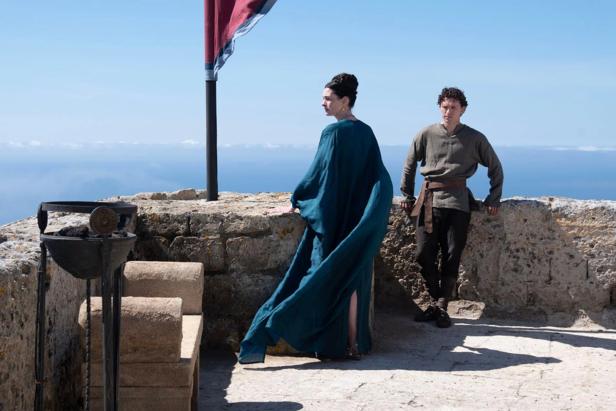 A woman in a long blue dress and a man in simple clothing stand on a stone terrace in front of the sea.