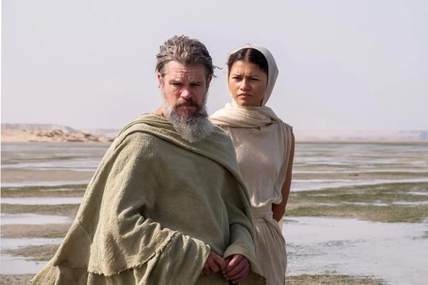 A bearded man and a woman with headscarves in bright, antique robes stand in a wide, flat desert landscape.