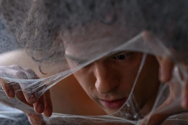 A young man with curly hair looks intently through a web of sticky, spider-web-like threads.