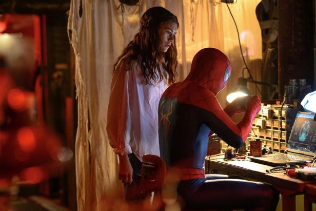 Spider-Man sits at a laptop and works on technical equipment while MJ stands nearby and watches him intently.