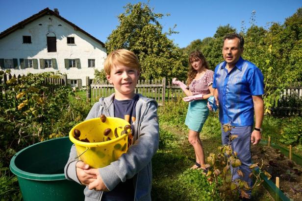 A boy holds a bucket with collected snails in the garden, two adults stand behind him.