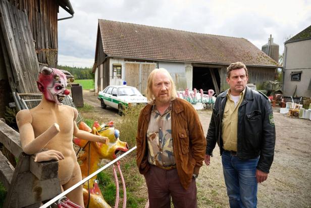 Three men stand in front of a farm, on the left a scary figure and a colorful wooden figure, in the background a police car.