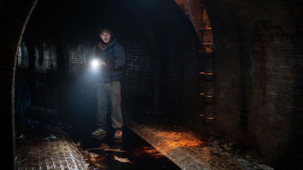 Person in dark tunnel with flashlight, on wet ground and in front of sooty brick walls.