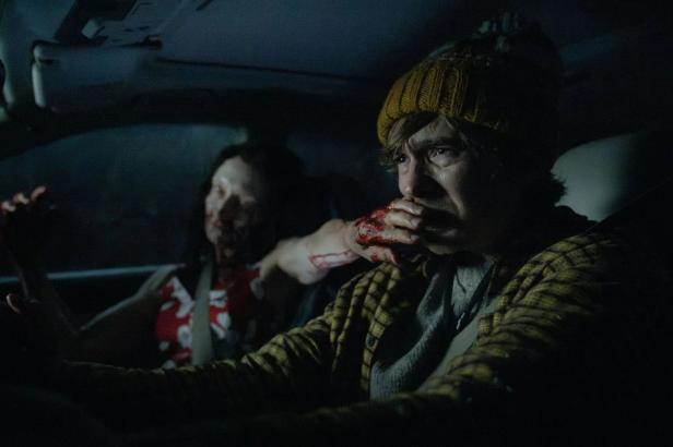 A man in a wool hat drives a car while a bloodied woman in the passenger seat puts her hand over his mouth.
