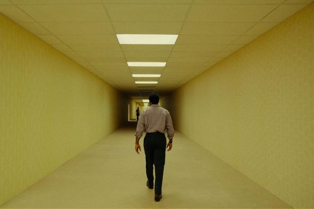 A man walks down a long, yellow hallway with fluorescent lights on the ceiling toward another person in the distance.