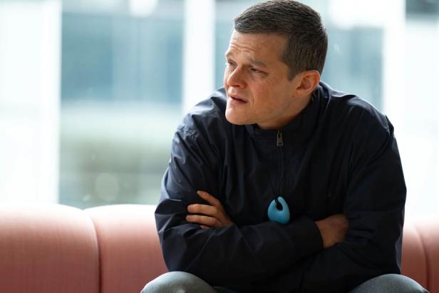 A man with his arms crossed sits on a sofa and looks thoughtfully to the side.