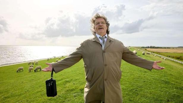 Hape Kerkeling as Horst Schlämmer stands with his arms outstretched in a sheep meadow by the sea