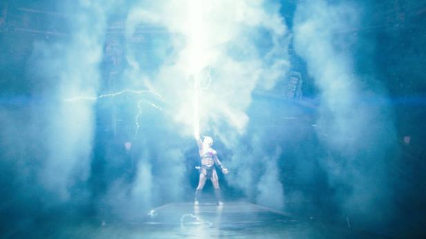 He-Man stands on a stage and lets a bright flash of lightning rise from his sword into the blue mist.