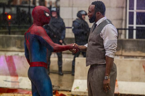 Spider-Man shakes hands with a man in a vest and shirt in the rain, while police officers in riot gear stand in the background.