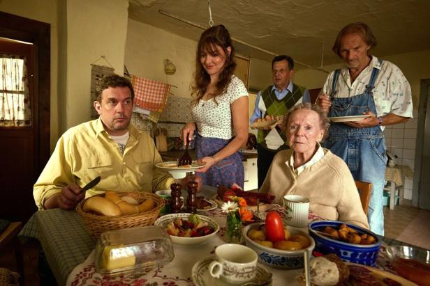 Several people sit and stand at a richly laid table in a kitchen while a woman serves food.