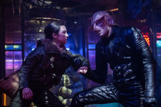 A young woman and an alien in black armor exchange a fist salute in a dark, neon-lit environment.