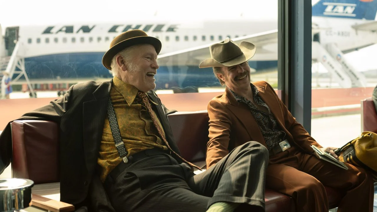 In the first scene at the airport, John Malkovich and Sam Rockwell go all out when it comes to dry humor.